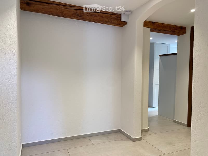 Empty hallway with white textured walls, tiled floor, and exposed wooden beams on the ceiling. A doorway leads to another room with a visible refrigerator.