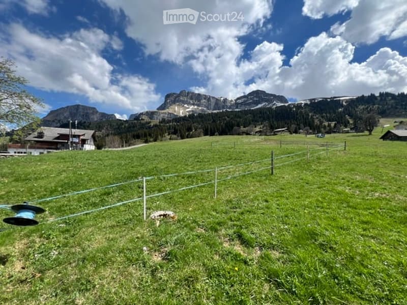 A serene, green meadow with a white fence in the foreground, surrounded by trees and mountains under a partly cloudy sky. A few houses are visible in the distance.