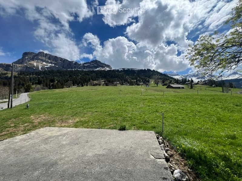A wide, open green field with scattered trees and a few small structures, surrounded by mountains under a partly cloudy sky.