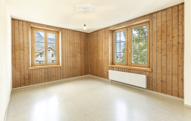 Empty room with wooden panel walls, large windows, and a radiator. The space is unfurnished with a clean, minimalist design.