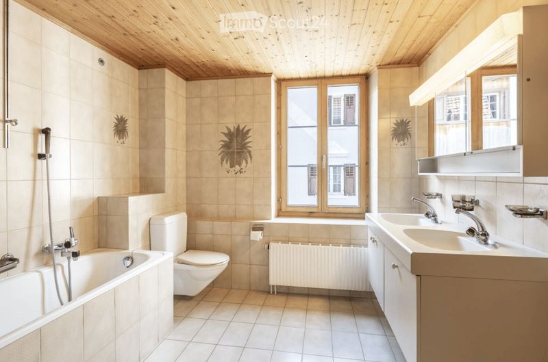 Bright bathroom with beige tiles, wooden ceiling, bathtub, toilet, double sink, and large window offering a view of the street.