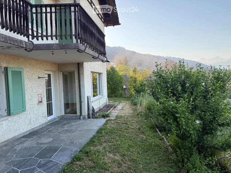 A pathway leads to a house with a balcony, green shutters, and a view of mountains in the background.