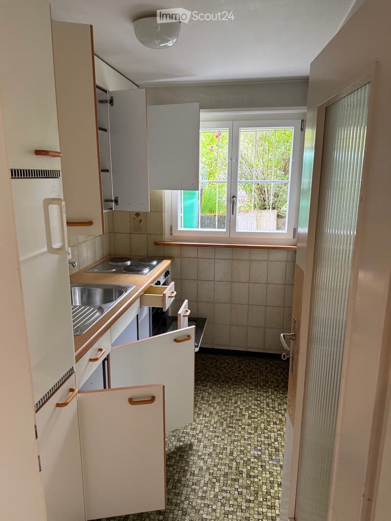 The kitchen features white cabinets with wooden handles, a sink, a stove, and an oven. The floor has a patterned tile design, and there is a window with a view of greenery outside.