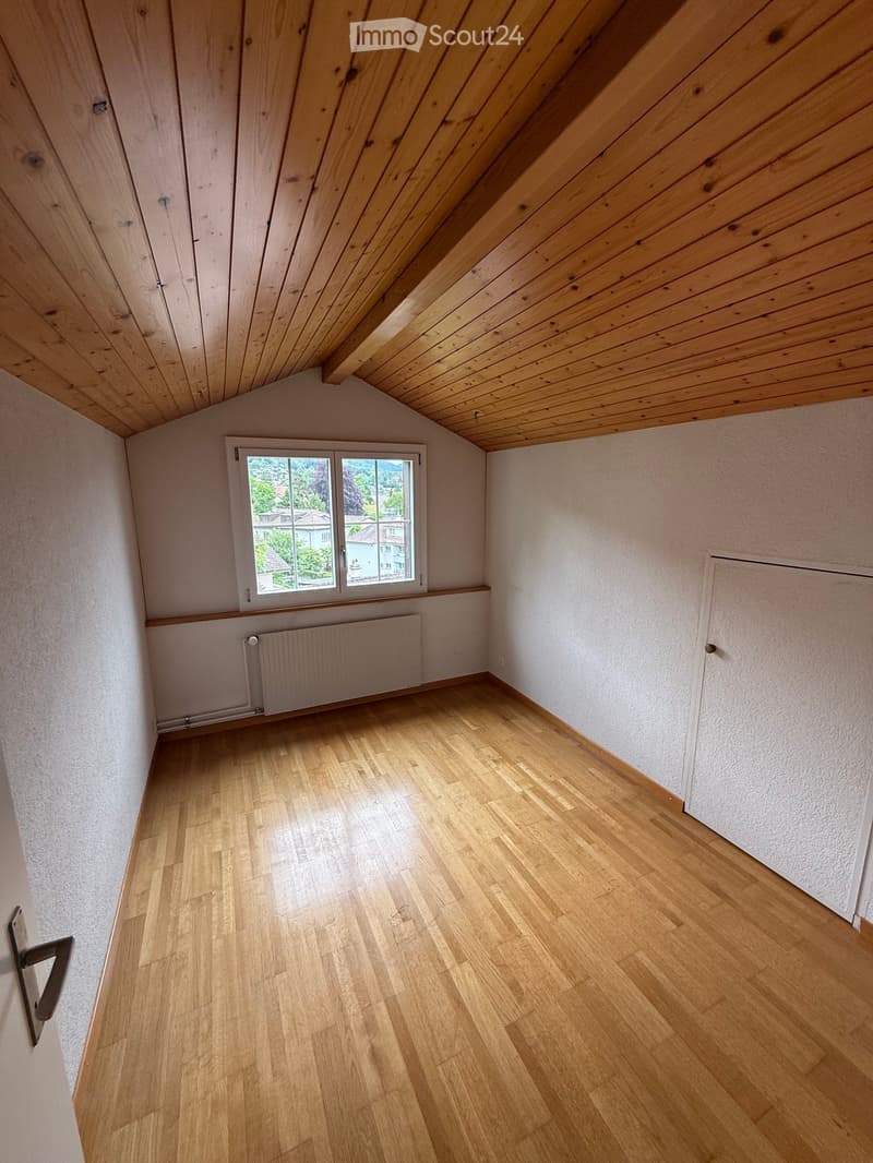 Empty room with wooden flooring, white walls, and a window offering a view of the outside. The ceiling has wooden paneling, and there is a radiator below the window.