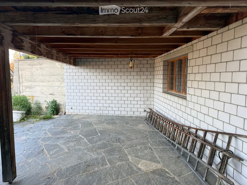 Covered outdoor area with stone flooring, white brick walls, wooden ceiling beams, and a small window with wooden frame.