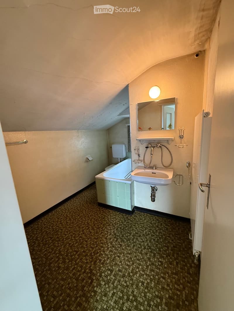 Small bathroom with a sink, bathtub, toilet, mirror, and light fixture. The room has a slanted ceiling and patterned carpet flooring.