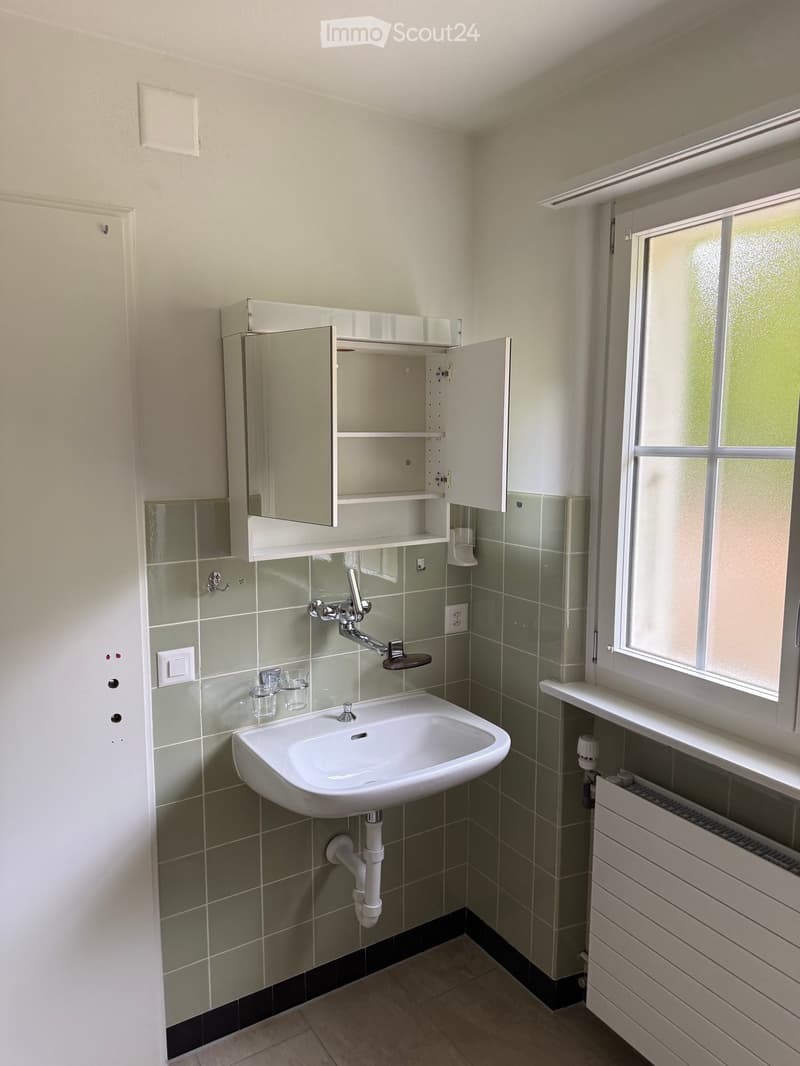 Modern bathroom with a white sink, mirror cabinet, tiled walls, and a window providing natural light.