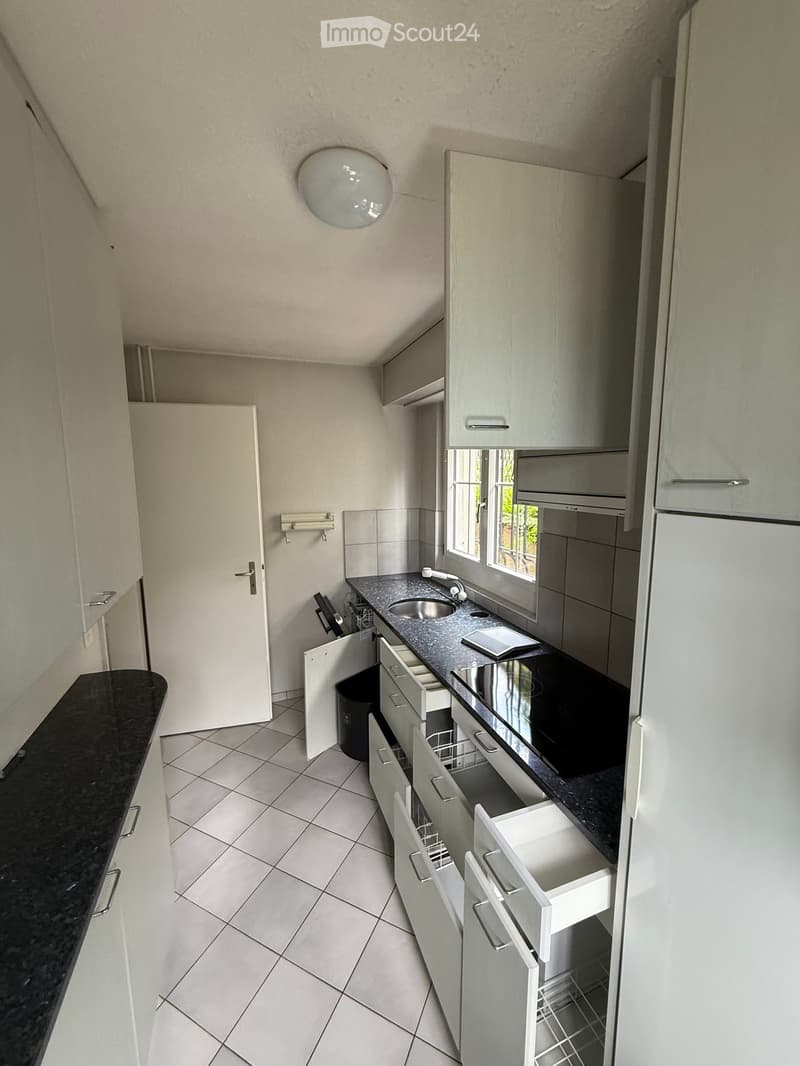 Modern kitchen with grey cabinets, black countertops, and a stainless steel sink. The space features tiled flooring and a window providing natural light.