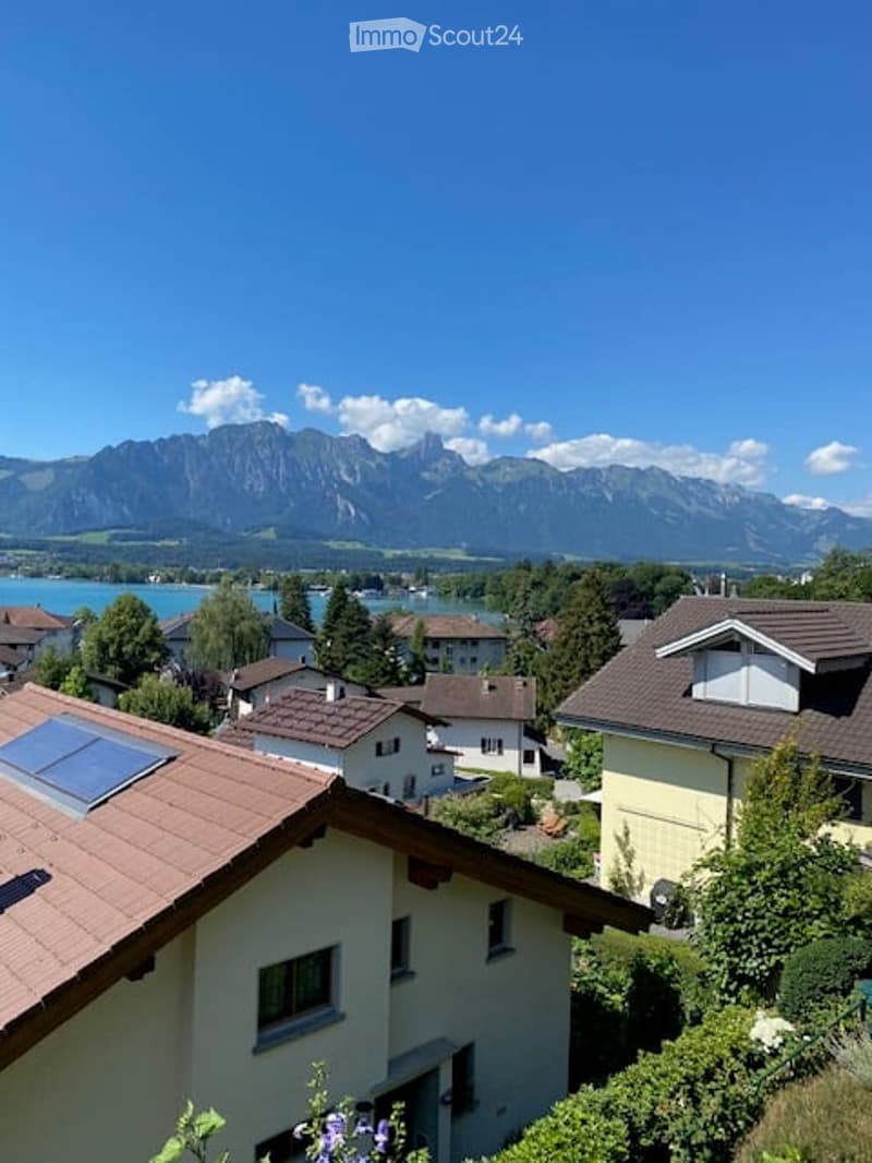 Residential houses with red-tiled roofs and solar panels, surrounded by greenery and overlooking a scenic mountain range and lake.