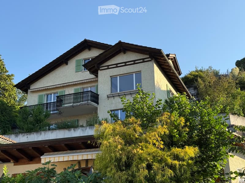 Two-story house with light-colored facade, green shutters, and a balcony on the upper level, surrounded by lush greenery.