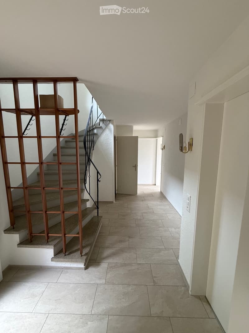Empty hallway with tiled floor, white walls, and a staircase leading upwards with wooden railings and a black handrail.