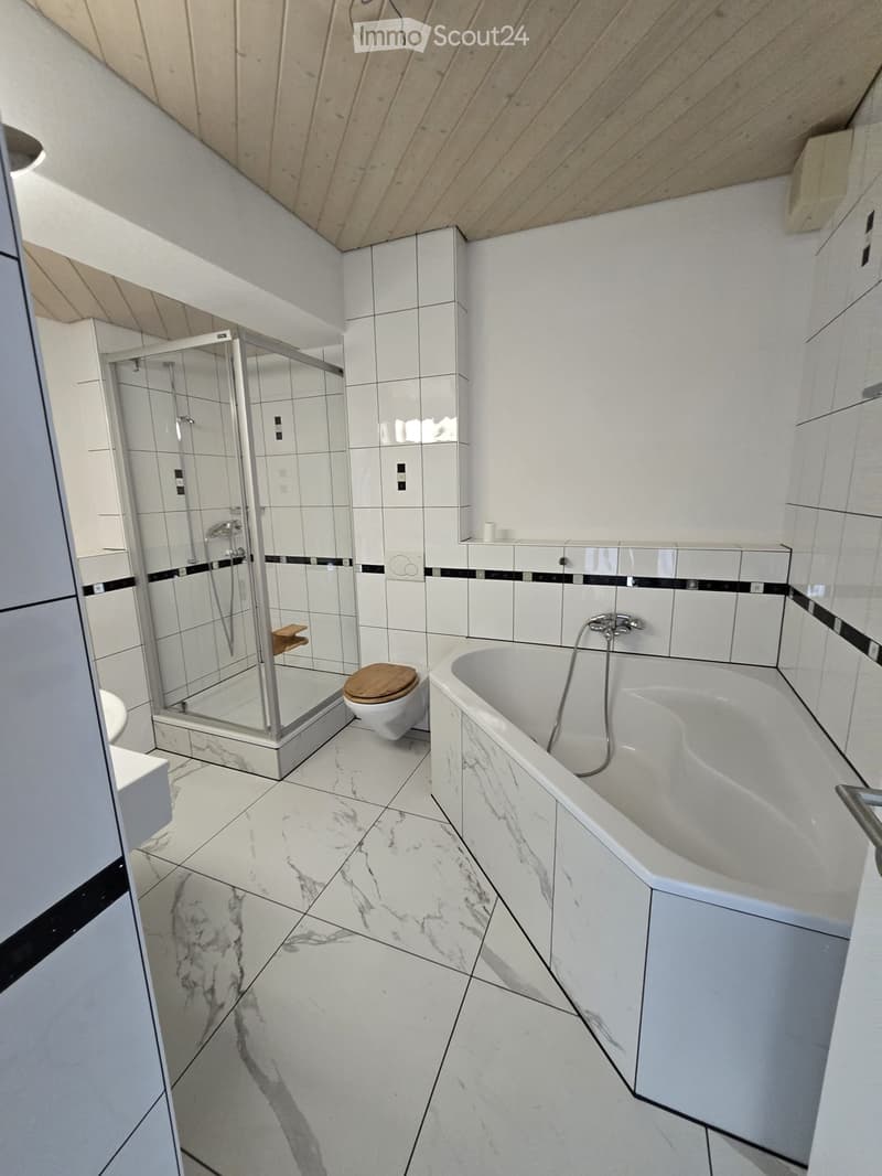 Modern bathroom with white marble tiles, a bathtub, a toilet, and a glass-enclosed shower. The shower has a wooden shelf inside.