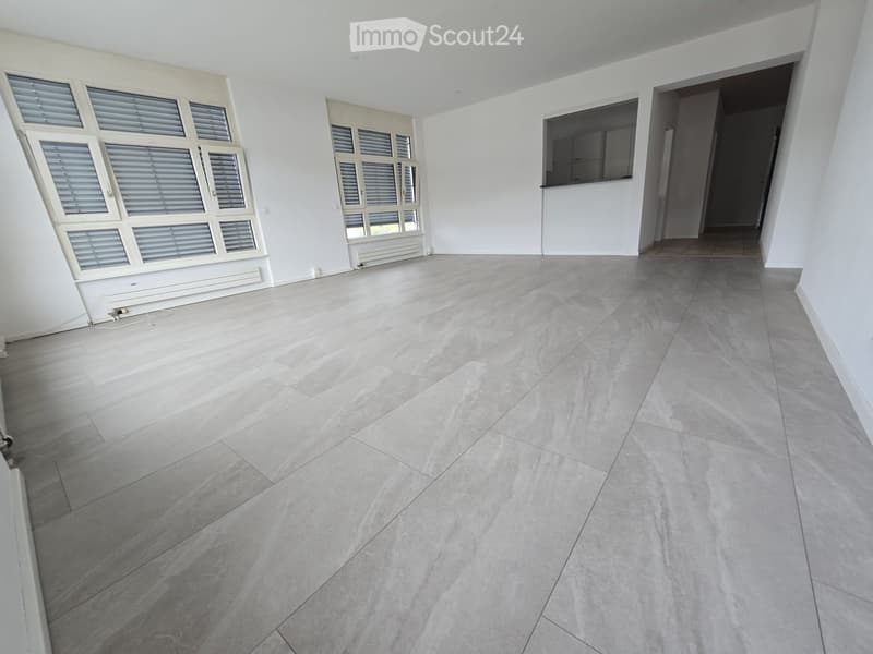 Empty room with light grey tiled floor, white walls, large windows with blinds, and a doorway leading to another space.