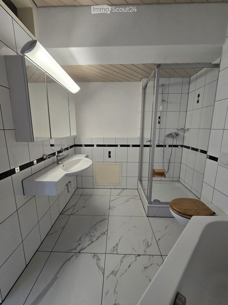 Modern bathroom with marble flooring, white tiles, a sink, toilet with wooden seat, and a glass-enclosed shower area.