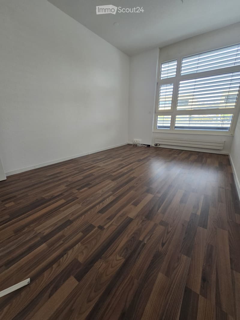 Empty room with polished wooden flooring, white walls, and large windows with blinds. No furniture or equipment present.