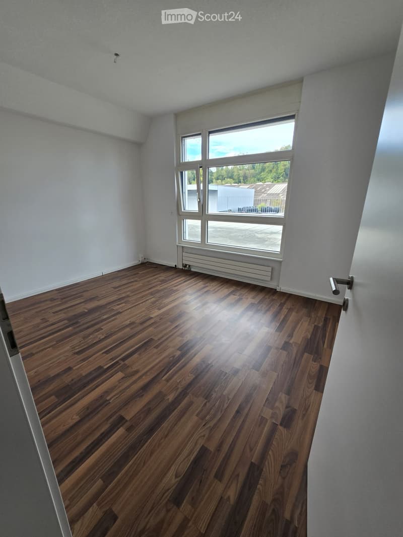 Empty room with wooden flooring, white walls, and large windows offering a view of the outside area.