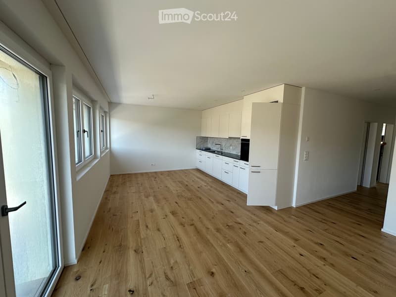 Empty room with wooden floor, white walls, kitchen with white cabinets, sink, faucet, and oven, large windows, sliding door
