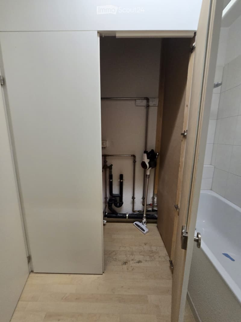 Empty room with exposed pipes and a vacuum cleaner, adjacent to a tiled bathroom with a bathtub.