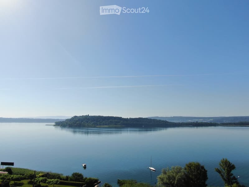 A serene lakeside view with calm water, distant tree-lined shore, and a few small boats anchored near the bank.