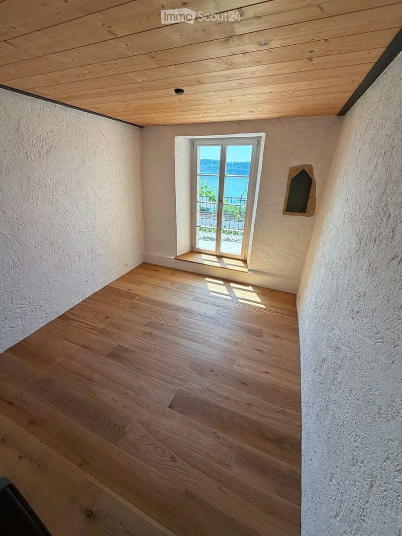 Empty room with wooden flooring, white walls, and a window offering a scenic view of the water and greenery.