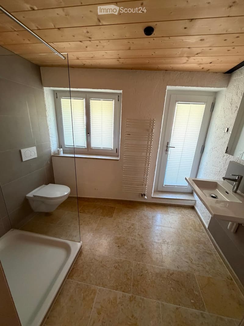 Modern bathroom with tiled floors, a bathtub, toilet, sink, and a radiator. Features large windows with blinds and a glass shower enclosure.