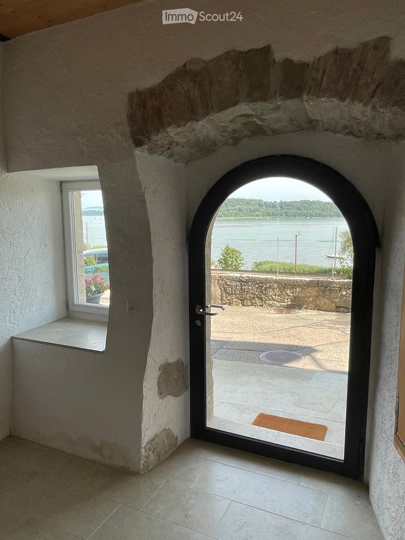 A hallway with a large arched doorway leading to an outdoor area with a view of a river and boats. The hallway has exposed stone walls and a tiled floor.