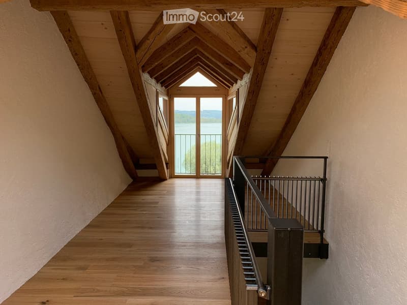 Spacious hallway with wooden floor, exposed wooden beams, and large windows leading to a balcony overlooking a lake.