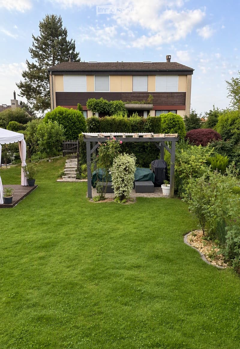 Well-maintained garden with a large lawn, a covered patio area with furniture, a pergola, and a house in the background.