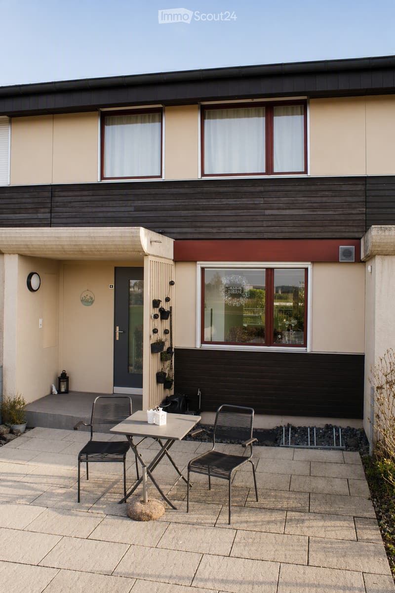 Outdoor patio area with a small table and two chairs, attached to a modern house with large windows and a wooden facade.