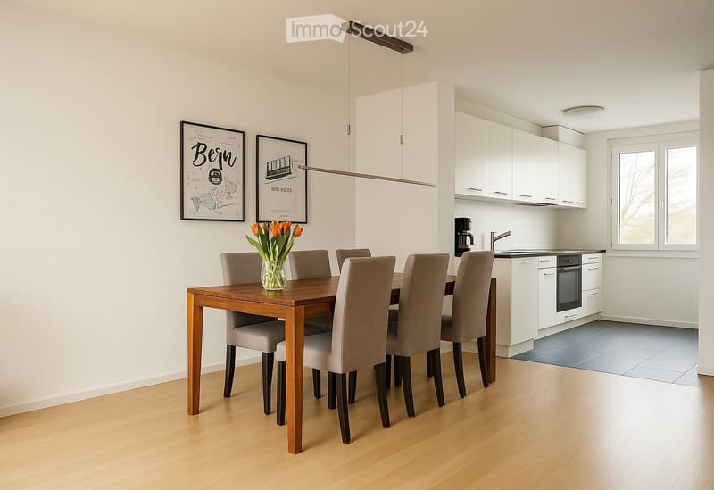 Open-plan dining and kitchen area with a wooden dining table, six chairs, a vase with flowers, white kitchen cabinets, a coffee maker, and framed artwork on the wall.