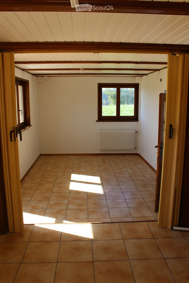 Empty room with tiled floor, wooden beams on the ceiling, and large windows allowing natural light to enter. The walls are painted in a light color, and there is a radiator on one wall.