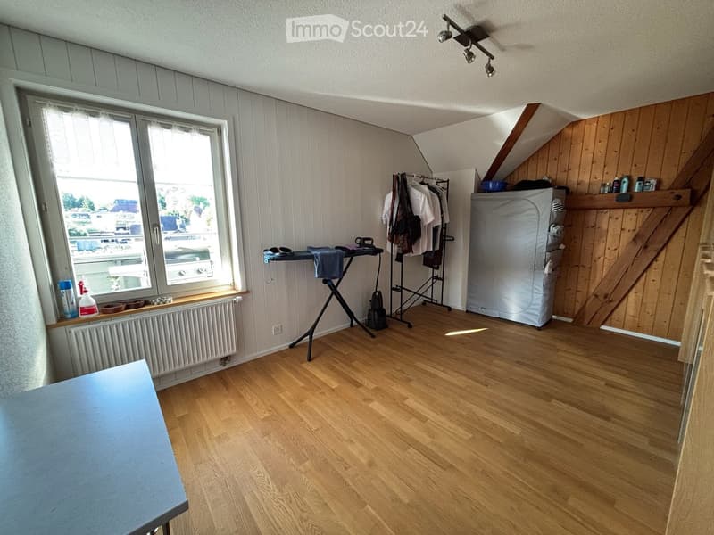 A small, furnished room with wooden flooring, a large window, a radiator, a clothing rack, an ironing board, and a refrigerator.