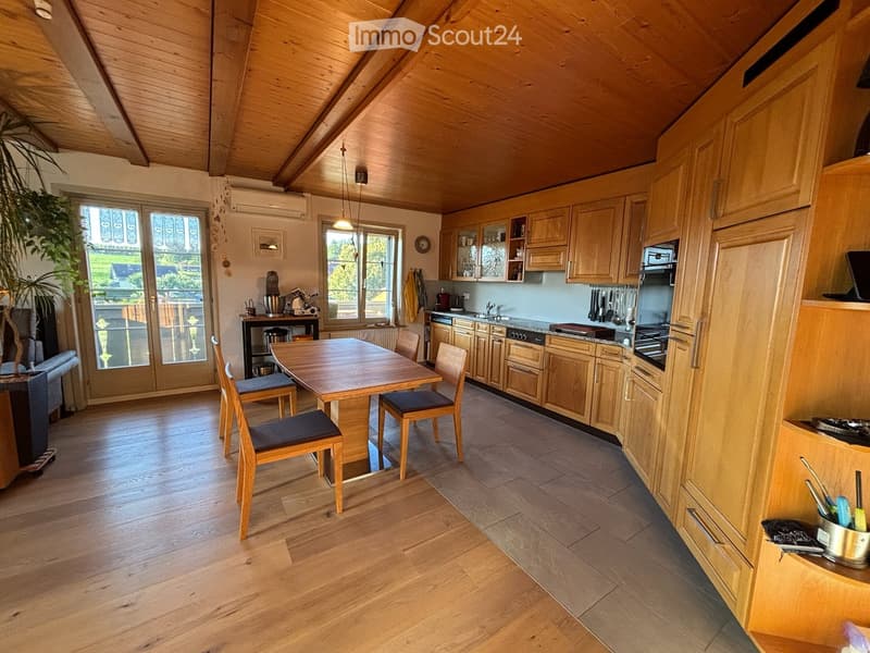 Bright kitchen with wooden cabinets, a central dining table with four chairs, and modern appliances including a sink, oven, and microwave. The space features wooden flooring and large windows with a view of the outdoors.