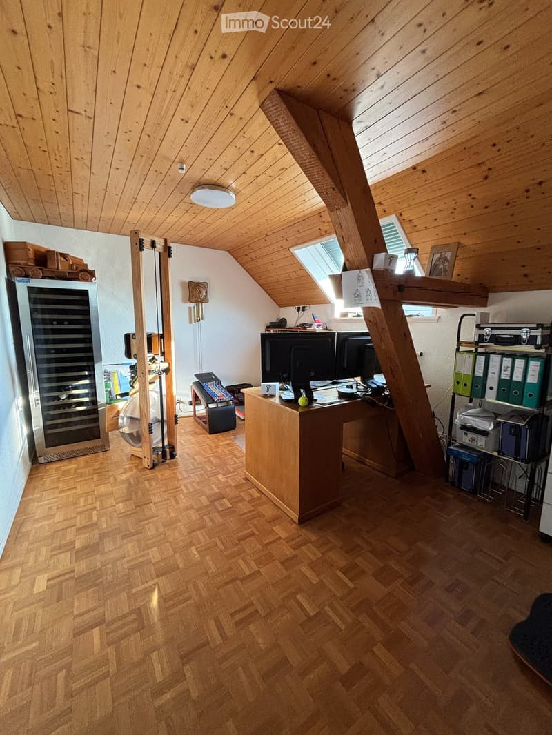 Office space with wooden herringbone flooring, slanted wooden ceiling, skylight, and various office equipment including a desk, computer, wine cooler, and shelves.