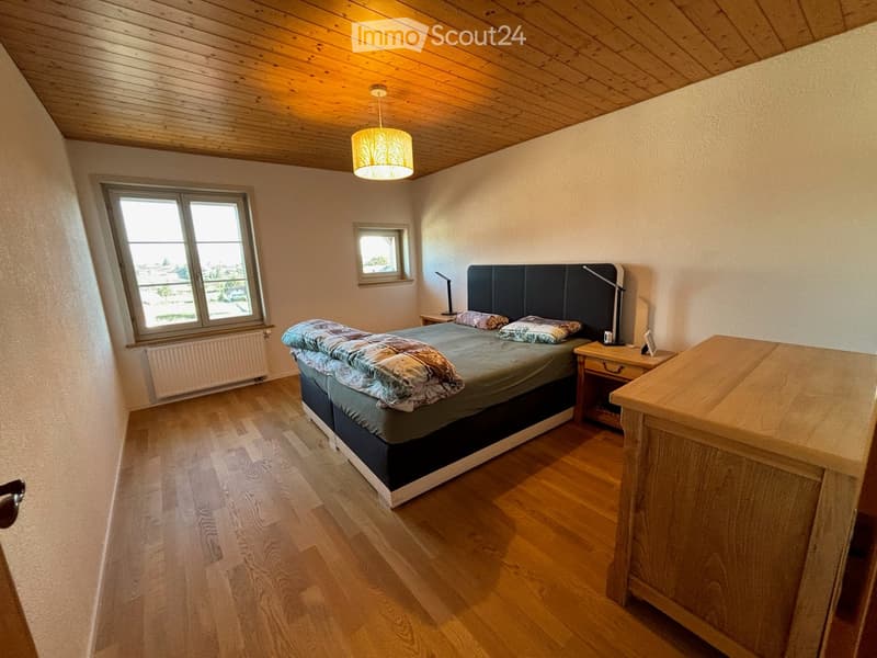 Cozy bedroom with wooden flooring, slanted wooden ceiling, large window, and a neatly made bed with pillows and blankets.