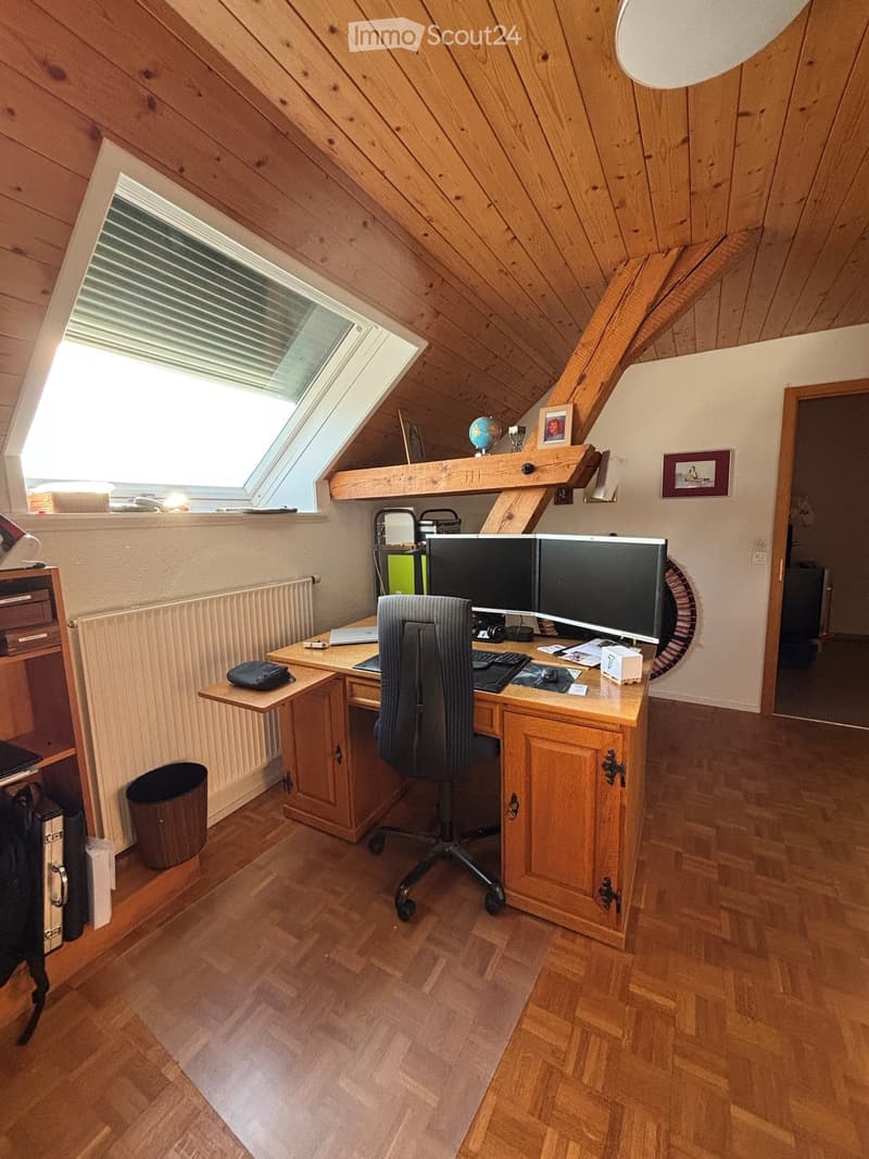Cozy attic office with wooden walls and slanted ceiling, featuring a large skylight window, a wooden desk with dual monitors, ergonomic chair, laptop, keyboard, mouse, globe, picture frame, and radiator.