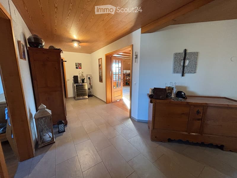 The image shows a well-lit hallway with wooden beams on the ceiling, tiled flooring, and several pieces of furniture including a wooden cabinet, a small refrigerator, a table with chairs, and decorative lanterns. There are also picture frames on the walls