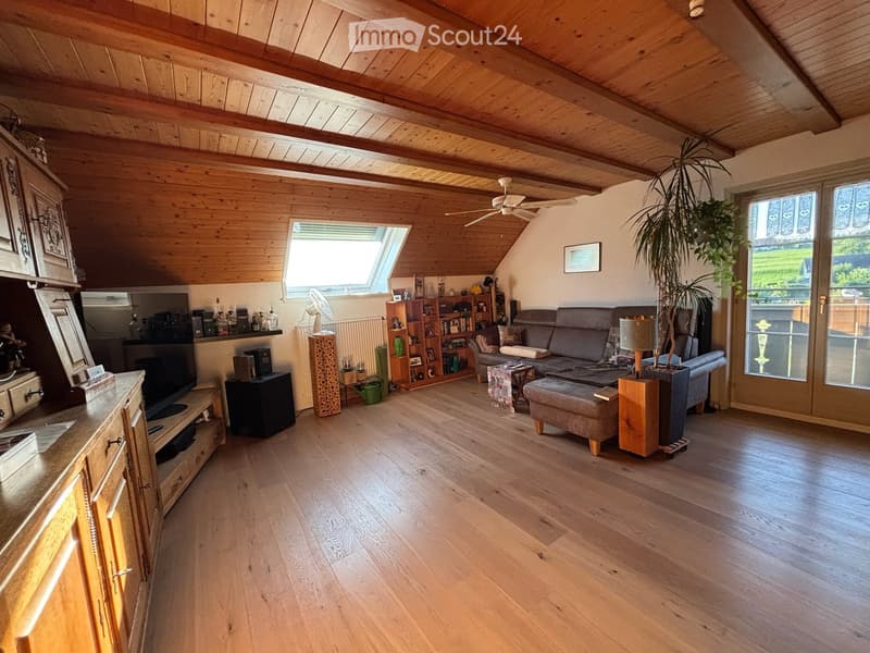 Bright living room with wooden floors and exposed wooden beams, featuring a sofa, ottoman, coffee table, bookshelf, and a large window letting in natural light.