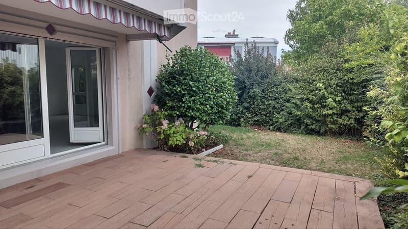 Brick patio area adjacent to a house with striped awning, glass sliding doors, and surrounding greenery including bushes and small plants.