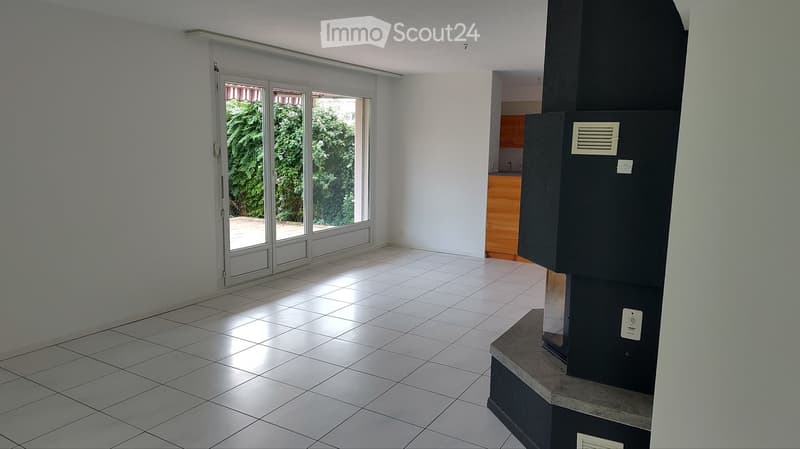 Empty room with tiled floor, white walls, large sliding glass doors leading to an outdoor area, and a small kitchen area visible in the background.