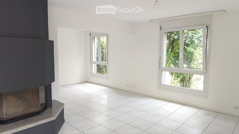 Empty room with white tiled floor, white walls, and two windows showing greenery outside. A fireplace is present on the left side.