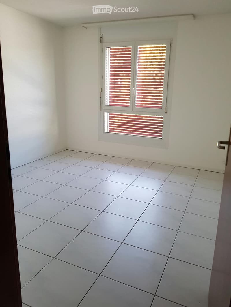 Empty room with white tiled floor and white walls, featuring a window with blinds and a door frame visible on the right side.