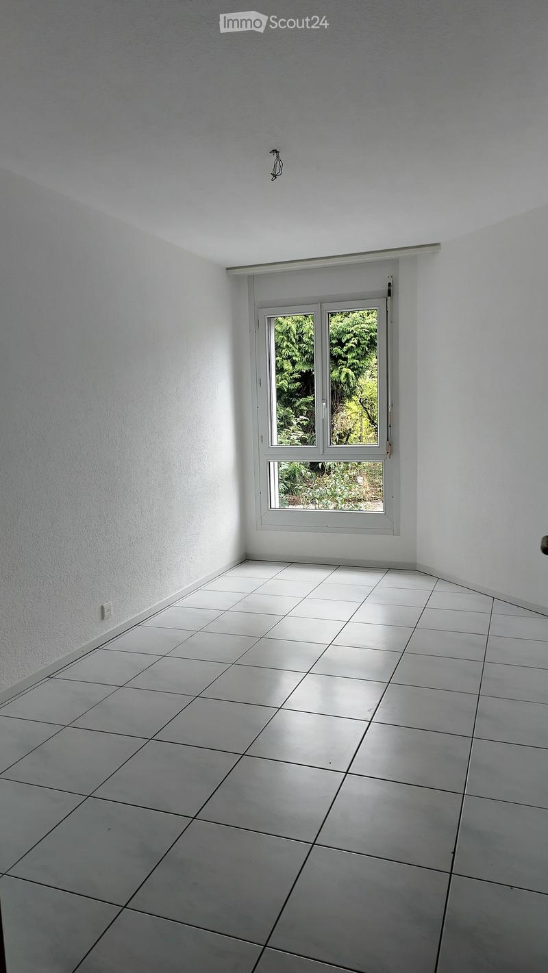 Empty room with white tiled floor and white walls, featuring a large window with a view of greenery outside.