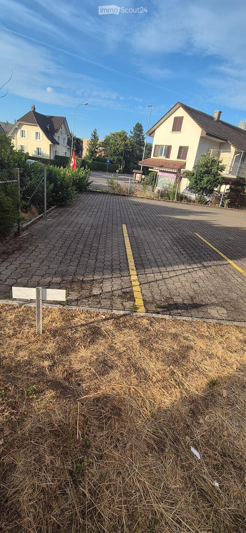 A paved driveway leading to a residential area with houses, greenery, and a street sign.