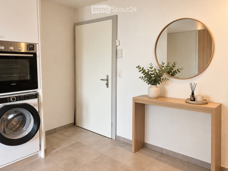 A small, modern hallway with a washing machine and oven on the left, a wooden console table with a vase and candle on the right, and a round mirror above the table.
