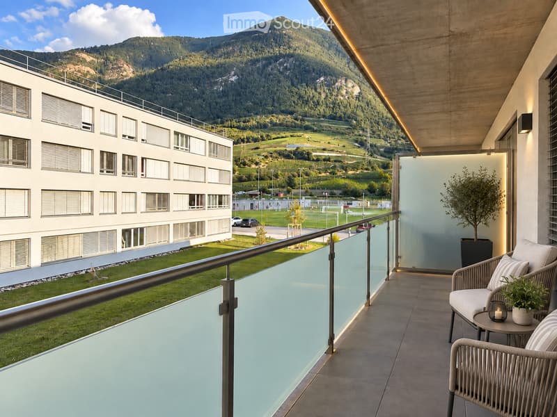 Modern balcony with a view of a mountain range, featuring a glass railing, a wicker chair, a small table with a candle, and potted plants.