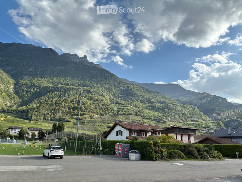A small house with a red-tiled roof and white walls, situated in a scenic valley with green hills and mountains in the background. A white car is parked in the foreground, and a fenced sports field is visible to the left.