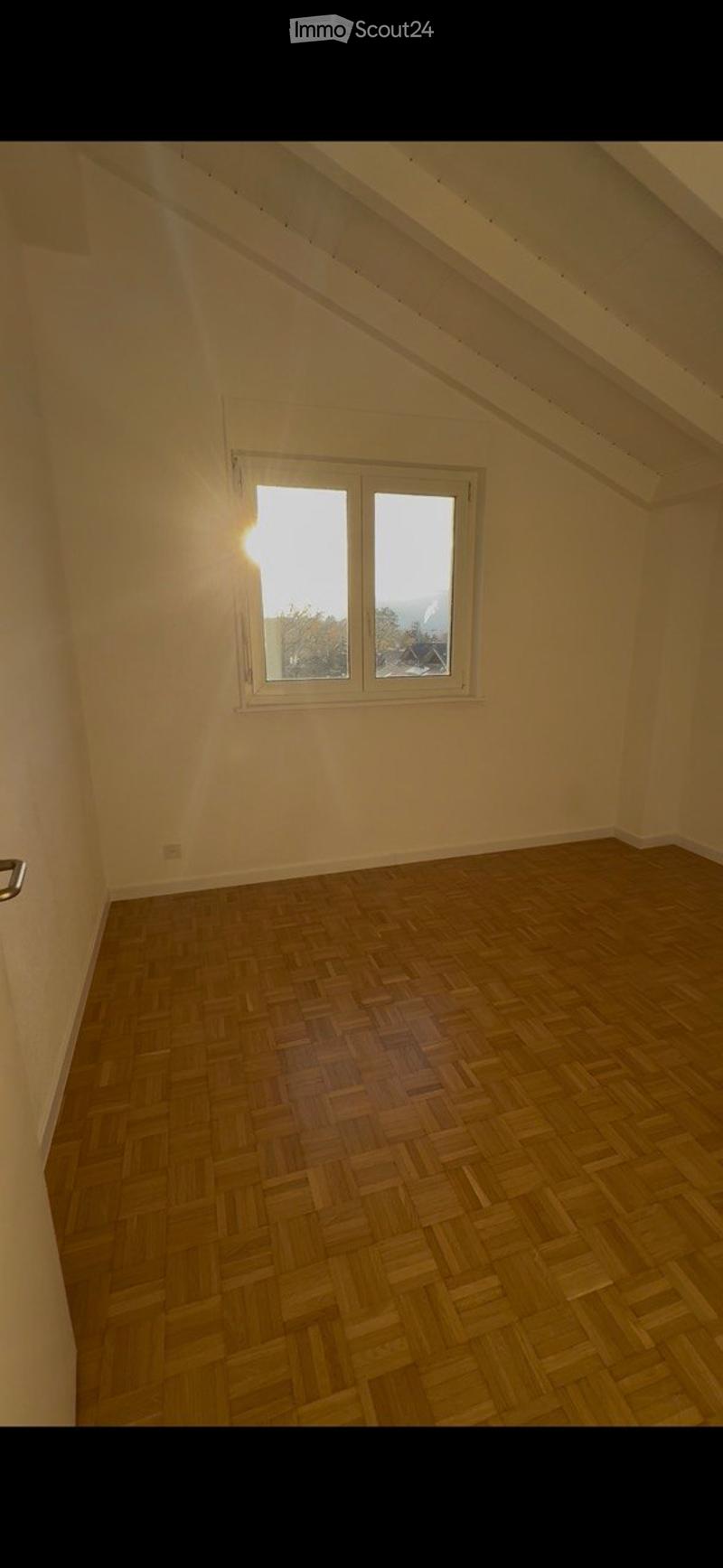 Empty attic room with wooden herringbone flooring, white walls, and a single window letting in natural light.