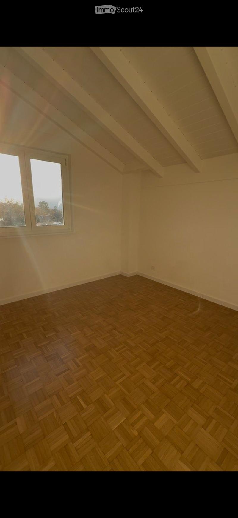 Empty attic room with wooden herringbone flooring, white walls, and exposed wooden beams on the ceiling. A single window allows natural light to enter.