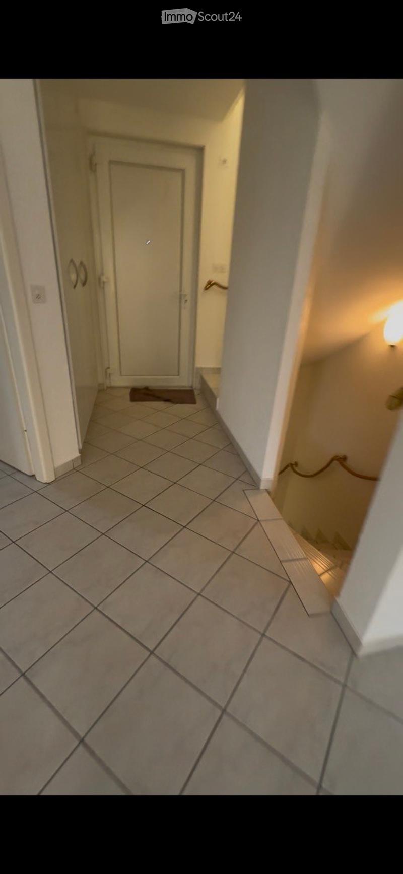 A narrow hallway with tiled flooring, white walls, and a closed door at the end. A small rug is placed in front of the door, and a light fixture is mounted on the right wall.